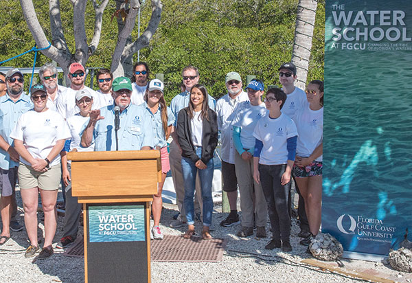 Man in blue shirt and green baseball camp standing at podium outdoors while people stand behind him