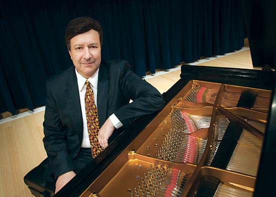 MAn wearaing dark suit and white shirt sitting at a piano looking at camera