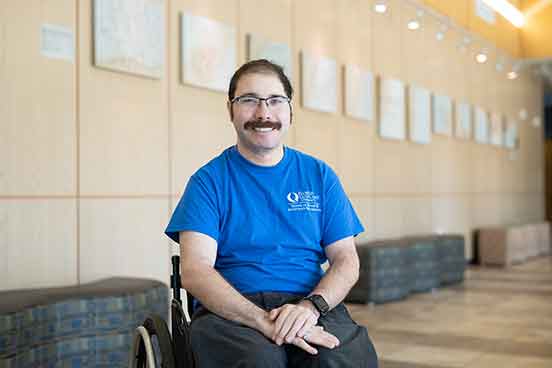 A man with glasses and a moustache in a blue, short-sleeved shirt sits in a wheelchair